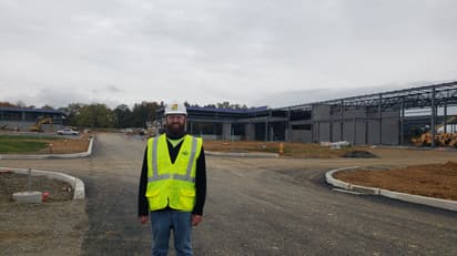 A young man standing at a jobsite in a construction vest, safety glasses, and a hardhat smiles at the camera.
